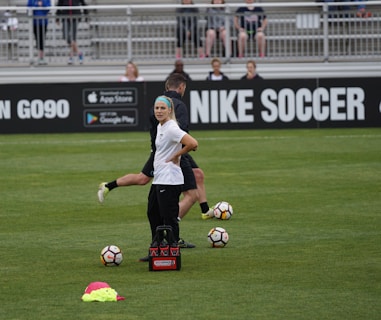 A person in sports attire stands on a soccer field, surrounded by several soccer balls and a carrier with bottles. The field is bordered by advertisements for Nike Soccer and app stores. Several people sit in bleachers in the background.