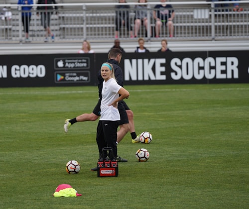 A person in sports attire stands on a soccer field, surrounded by several soccer balls and a carrier with bottles. The field is bordered by advertisements for Nike Soccer and app stores. Several people sit in bleachers in the background.