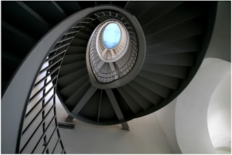 A spiral staircase with a central circular opening leading to a small skylight at the top. The staircase is composed of dark steps contrasted against a lighter background, providing a striking visual effect. The architectural structure creates a dynamic sense of movement and depth.