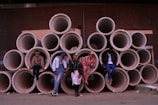 A group of five individuals pose confidently in front of a pile of large, stacked concrete pipes. Each person is dressed in colorful, eclectic clothing, expressing a unique and bold sense of style. The backdrop features a dark brick wall that adds an industrial urban aesthetic to the scene.