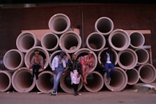 A group of five individuals pose confidently in front of a pile of large, stacked concrete pipes. Each person is dressed in colorful, eclectic clothing, expressing a unique and bold sense of style. The backdrop features a dark brick wall that adds an industrial urban aesthetic to the scene.