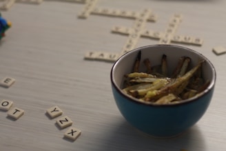 A bowl filled with seasoned fries sits on a table with scattered Scrabble tiles. The scene suggests a casual and relaxed setting that combines dining with a word game. The Scrabble board has partially completed words made from the beige tiles.