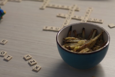 A bowl filled with seasoned fries sits on a table with scattered Scrabble tiles. The scene suggests a casual and relaxed setting that combines dining with a word game. The Scrabble board has partially completed words made from the beige tiles.