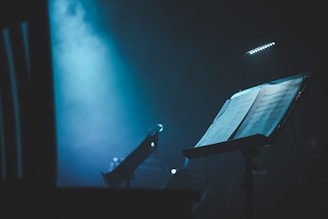 A dimly lit music stand holds sheets of music, illuminated by a small clip-on light. In the background, there is a saxophone resting on a chair, with a hazy blue atmosphere enhanced by stage lighting.