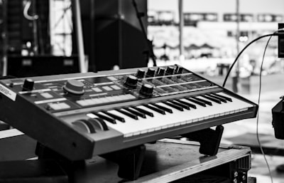 A black and white image featuring a synthesizer keyboard positioned on a stand outdoors. The background appears to be a stage setup, likely prepared for a musical performance. Various control knobs and buttons are visible on the synthesizer, suggesting a range of sound modulation options.