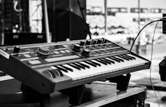 A black and white image featuring a synthesizer keyboard positioned on a stand outdoors. The background appears to be a stage setup, likely prepared for a musical performance. Various control knobs and buttons are visible on the synthesizer, suggesting a range of sound modulation options.