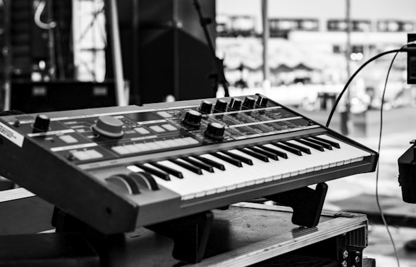 A black and white image featuring a synthesizer keyboard positioned on a stand outdoors. The background appears to be a stage setup, likely prepared for a musical performance. Various control knobs and buttons are visible on the synthesizer, suggesting a range of sound modulation options.