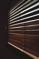 A close-up view of wooden blinds partially closed, casting shadows and allowing diffused light to pass through. The slats are positioned diagonally, creating an interplay of light and shadow on their surface.