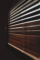 A close-up view of wooden blinds partially closed, casting shadows and allowing diffused light to pass through. The slats are positioned diagonally, creating an interplay of light and shadow on their surface.