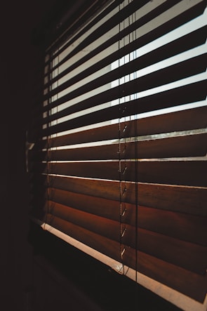 A close-up view of wooden blinds partially closed, casting shadows and allowing diffused light to pass through. The slats are positioned diagonally, creating an interplay of light and shadow on their surface.
