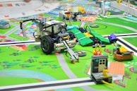 A detailed scene of a robotics competition setup with various LEGO robots and mechanized devices on a colorful mat. The mat depicts a city layout with roads, green areas, and small structures, giving it a playful and educational atmosphere. Several robots, equipped with wheels and sensors, are positioned at different points on the mat, engaged in various tasks. One notable robot in the foreground has an arm with a gripper or tool. Parts of the mat simulate different urban elements and activities, such as a small structure with flames and a figure, likely representing a house on fire scenario.