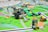 A detailed scene of a robotics competition setup with various LEGO robots and mechanized devices on a colorful mat. The mat depicts a city layout with roads, green areas, and small structures, giving it a playful and educational atmosphere. Several robots, equipped with wheels and sensors, are positioned at different points on the mat, engaged in various tasks. One notable robot in the foreground has an arm with a gripper or tool. Parts of the mat simulate different urban elements and activities, such as a small structure with flames and a figure, likely representing a house on fire scenario.