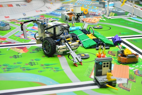 A detailed scene of a robotics competition setup with various LEGO robots and mechanized devices on a colorful mat. The mat depicts a city layout with roads, green areas, and small structures, giving it a playful and educational atmosphere. Several robots, equipped with wheels and sensors, are positioned at different points on the mat, engaged in various tasks. One notable robot in the foreground has an arm with a gripper or tool. Parts of the mat simulate different urban elements and activities, such as a small structure with flames and a figure, likely representing a house on fire scenario.