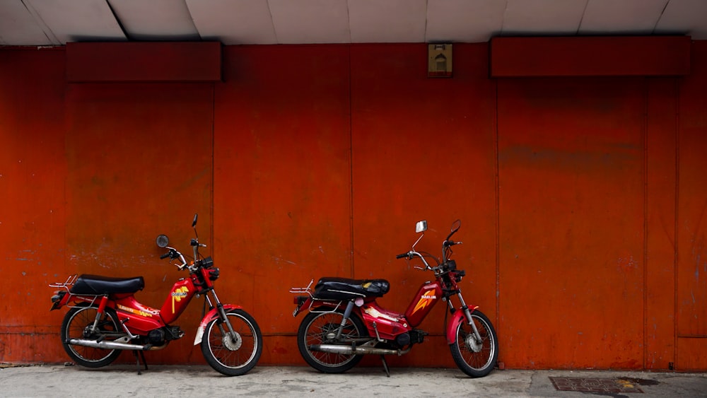 두 개의 빨간 페달 오토바이가 빨간 페인트 벽 옆에 주차되어 있습니다.