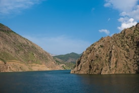 A serene landscape featuring a large body of water surrounded by rocky mountains with sparse greenery. The sky is clear with a few scattered clouds, highlighting the natural beauty of the scene.