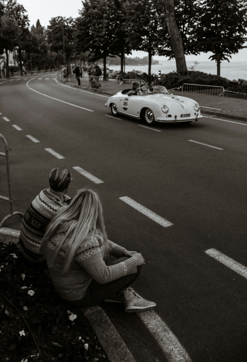grayscale photo of man and woman sitting on street facing white coupe
