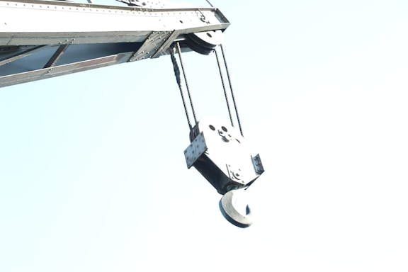 A close-up view of a crane's hook, showing metal cables and mechanical components against a bright, clear sky.