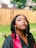A woman is outdoors wearing a black graduation gown with a red stole. She is looking upward, with long earrings and styled hair. The background features a wooden fence and a green lawn, suggesting a backyard setting.