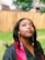 A woman is outdoors wearing a black graduation gown with a red stole. She is looking upward, with long earrings and styled hair. The background features a wooden fence and a green lawn, suggesting a backyard setting.