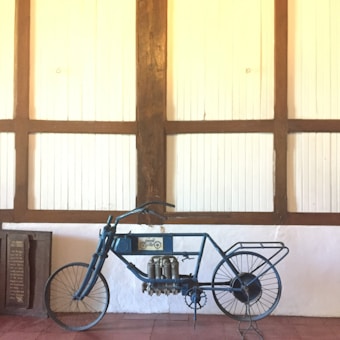 A vintage motorcycle with a unique and exposed mechanical design, showcasing intricate components and metal framework. Positioned against a wooden paneled wall, the scene evokes a sense of history and craftsmanship.