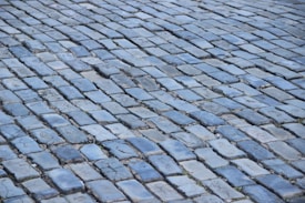 Cobblestone pavement with irregularly shaped, closely packed stones in various shades of blue and gray. Some stones have visible wear and cracks, with small patches of green moss or grass between them.