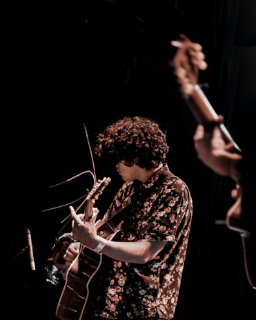 A musician with curly hair plays an acoustic guitar on stage. The person is wearing a floral patterned shirt and appears focused on the performance. The lighting is dim, creating a moody atmosphere suitable for a live music event.