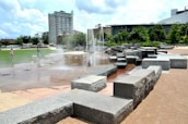 A modern outdoor urban space features a series of water fountains surrounded by large, geometric stone structures. In the background, there are green lawns and trees, and a high-rise building is visible. A person walks a dog and another person stands near the fountains.