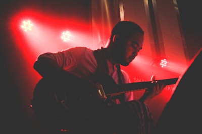 A musician is playing an electric guitar under dramatic red stage lighting. The silhouette highlights their focused expression and the intricate details of the guitar. Spotlights in the background create vibrant light effects, adding to the concert atmosphere.