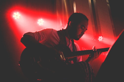 A musician is playing an electric guitar under dramatic red stage lighting. The silhouette highlights their focused expression and the intricate details of the guitar. Spotlights in the background create vibrant light effects, adding to the concert atmosphere.