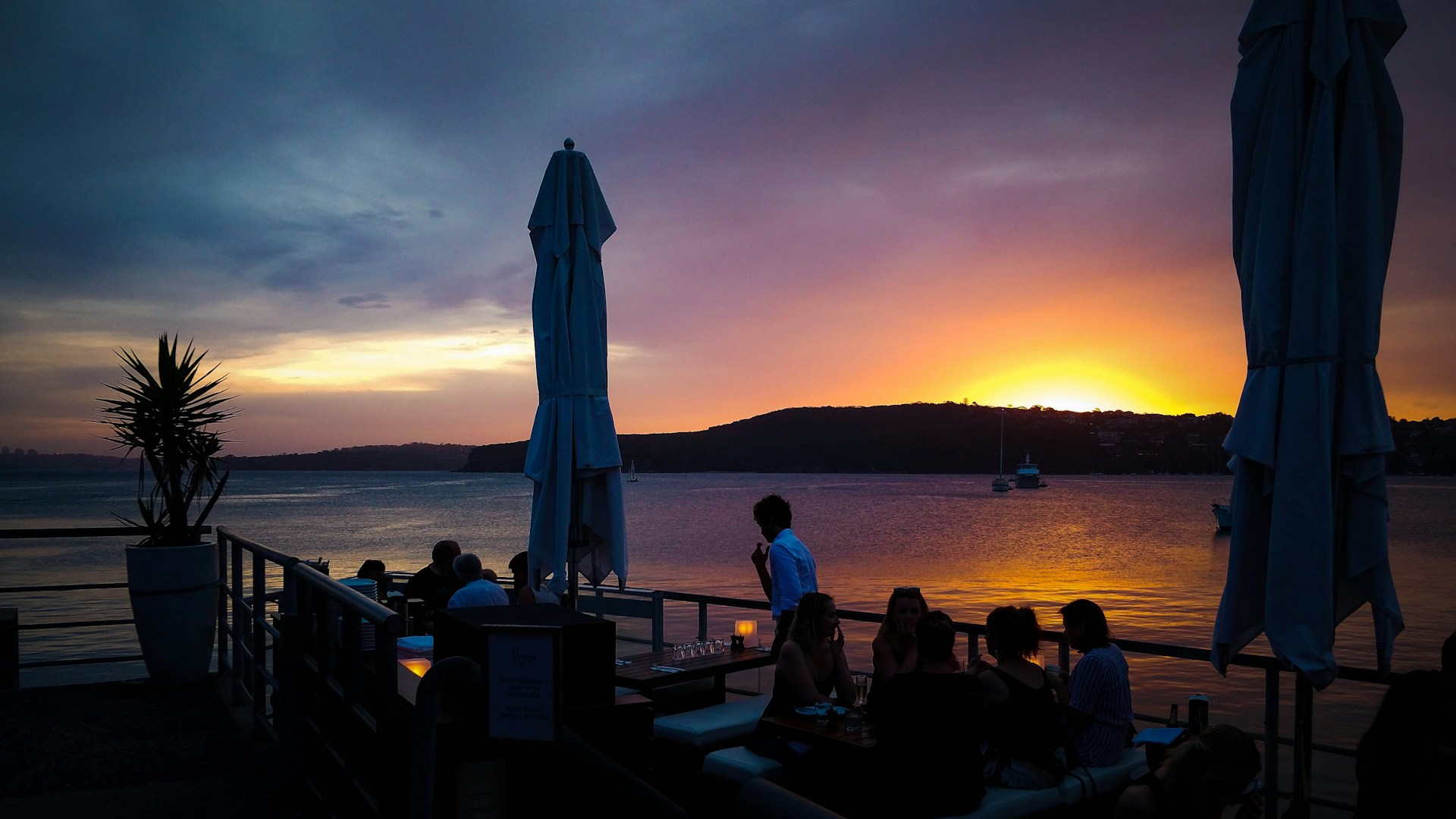 A vibrant sunset view over the river with outdoor tables of Las Brazzas Grill filled with happy diners enjoying mar and tierra dishes.