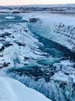 A stunning icy waterfall cascades down a rugged landscape, surrounded by snow-covered rocks and a vast expanse of ice. The water appears to flow through a deep gorge, creating a dramatic natural scene.