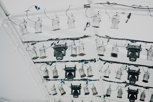 A collection of stage lights is mounted on a white metal truss with multiple black wires and cables attached. The lighting setup includes various types and sizes of spotlights and floodlights, some with adjustable panels.