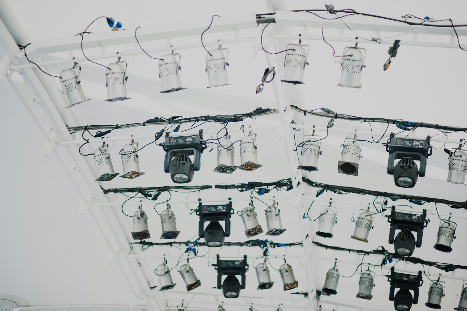 A collection of stage lights is mounted on a white metal truss with multiple black wires and cables attached. The lighting setup includes various types and sizes of spotlights and floodlights, some with adjustable panels.