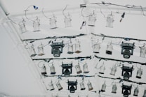 A collection of stage lights is mounted on a white metal truss with multiple black wires and cables attached. The lighting setup includes various types and sizes of spotlights and floodlights, some with adjustable panels.