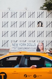 A yellow taxi with an advertisement on top passes in front of a wall covered in tally marks. A mural painted on the wall depicts a person behind bars, giving the impression of counting days. The background shows some greenery.
