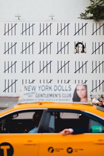 A yellow taxi with an advertisement on top passes in front of a wall covered in tally marks. A mural painted on the wall depicts a person behind bars, giving the impression of counting days. The background shows some greenery.