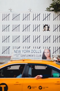 A yellow taxi with an advertisement on top passes in front of a wall covered in tally marks. A mural painted on the wall depicts a person behind bars, giving the impression of counting days. The background shows some greenery.