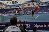 A tennis player stands on an indoor court holding a racket, ready to serve. The match is being watched by a sizeable audience, seated in blue and red stands. The umpire is visible on an elevated chair near the net. The court is well-lit, and various sponsors’ banners are displayed around the court edges.