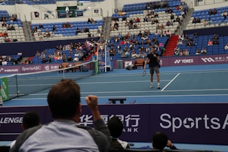 A tennis player stands on an indoor court holding a racket, ready to serve. The match is being watched by a sizeable audience, seated in blue and red stands. The umpire is visible on an elevated chair near the net. The court is well-lit, and various sponsors’ banners are displayed around the court edges.