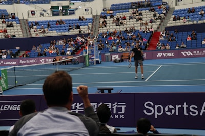 A tennis player stands on an indoor court holding a racket, ready to serve. The match is being watched by a sizeable audience, seated in blue and red stands. The umpire is visible on an elevated chair near the net. The court is well-lit, and various sponsors’ banners are displayed around the court edges.