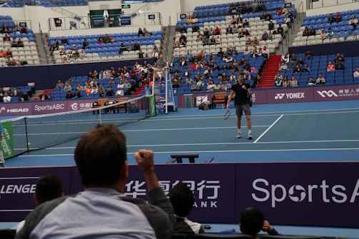 A tennis player stands on an indoor court holding a racket, ready to serve. The match is being watched by a sizeable audience, seated in blue and red stands. The umpire is visible on an elevated chair near the net. The court is well-lit, and various sponsors’ banners are displayed around the court edges.