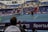 A tennis player stands on an indoor court holding a racket, ready to serve. The match is being watched by a sizeable audience, seated in blue and red stands. The umpire is visible on an elevated chair near the net. The court is well-lit, and various sponsors’ banners are displayed around the court edges.