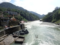 A flowing river surrounded by lush green mountains under a partially cloudy sky. A small temple structure with a dome and red flag is situated on the rocky banks, alongside several buildings. A suspended cable car chair hangs over the river, suggesting transport or scenic viewing.