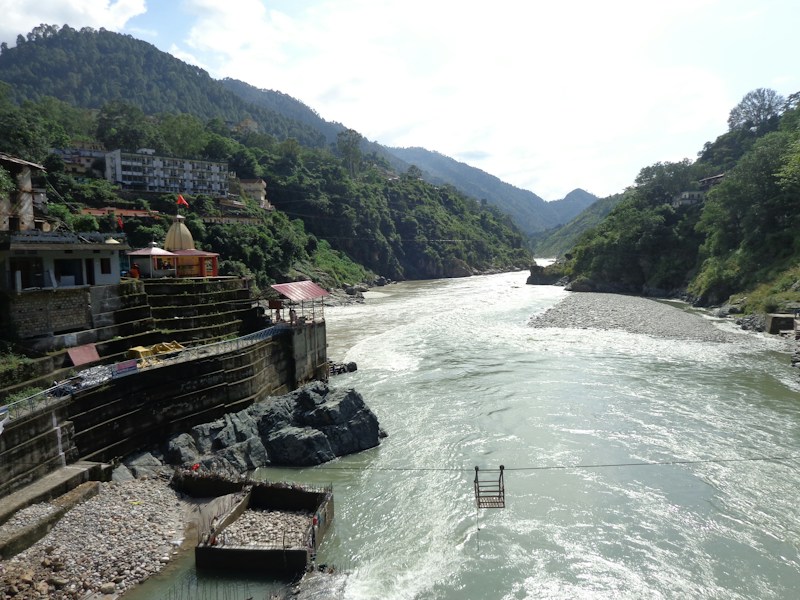 A flowing river surrounded by lush green mountains under a partially cloudy sky. A small temple structure with a dome and red flag is situated on the rocky banks, alongside several buildings. A suspended cable car chair hangs over the river, suggesting transport or scenic viewing.