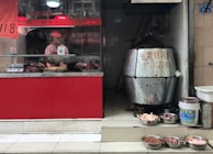 A small food shop with a display counter containing roasted meats. A person wearing a pink uniform and cap is behind the counter, preparing food. To the right, a large metal roasting oven is visible, next to various bowls filled with raw poultry parts on the tiled floor.