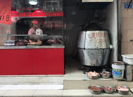 A small food shop with a display counter containing roasted meats. A person wearing a pink uniform and cap is behind the counter, preparing food. To the right, a large metal roasting oven is visible, next to various bowls filled with raw poultry parts on the tiled floor.