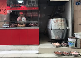 A small food shop with a display counter containing roasted meats. A person wearing a pink uniform and cap is behind the counter, preparing food. To the right, a large metal roasting oven is visible, next to various bowls filled with raw poultry parts on the tiled floor.