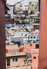 Colorful, densely packed houses line a hilly urban neighborhood, with buildings in various pastel shades of pink, green, and blue. The structures appear closely spaced, and the scene is intersected by numerous power lines crisscrossing the view.