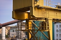 A large, industrial yellow crane structure stands prominently with its metal framework and ladder visible. In the background, various industrial buildings and structures, including cylindrical towers and a slanted brown pipe, are present. The area suggests an industrial setting, likely a factory or construction site, with a mix of modern and older building designs.