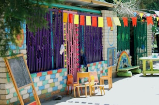A colorful outdoor setting features a wall painted with alternating blue, white, and pink bricks. Bright metal bars in purple and green cover the windows. A series of colorful triangular flags hang overhead. A small chalkboard and several wooden chairs are arranged near the wall. On the ground, a painted rainbow decorates the side of a small structure next to a circular green table.